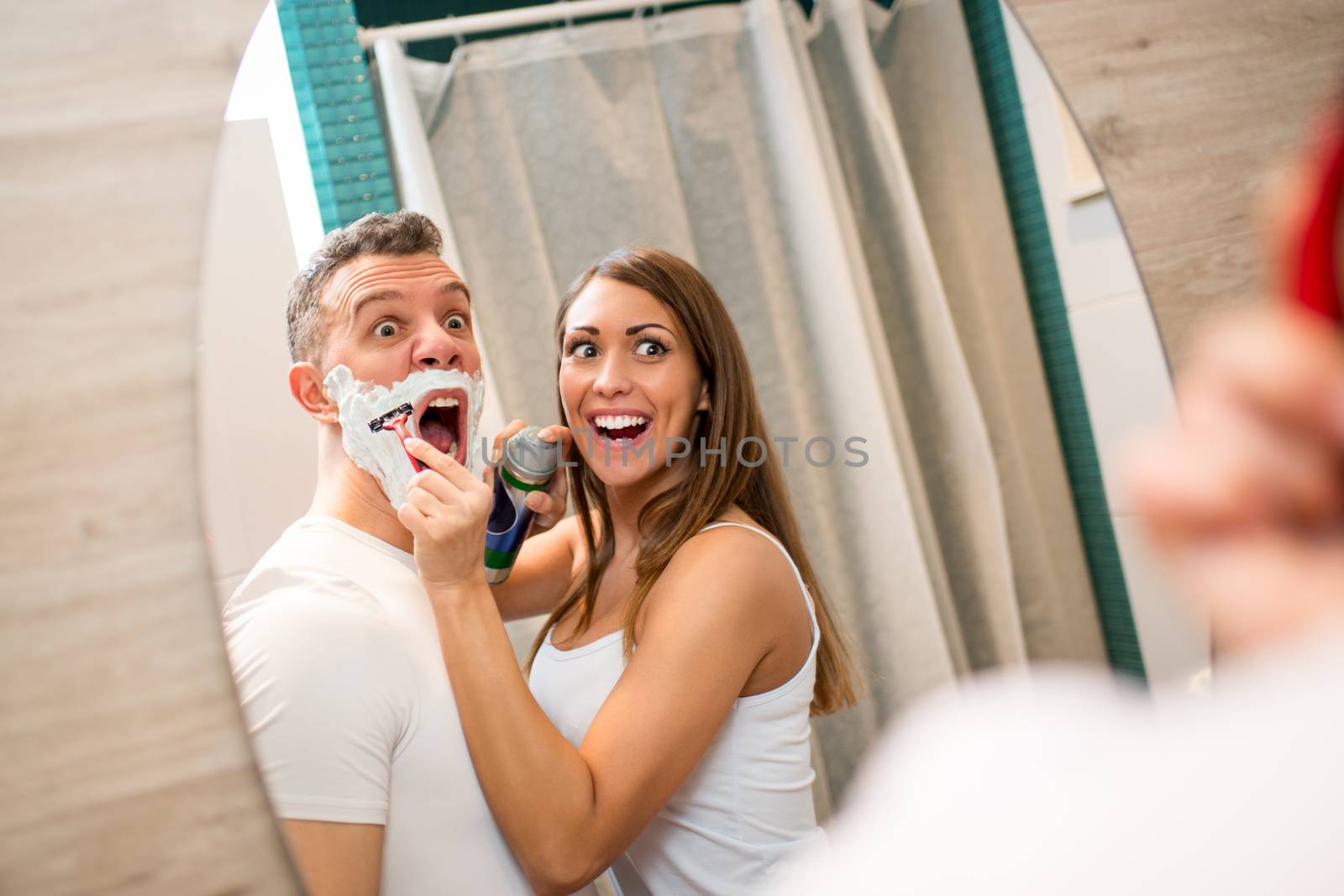 Portrait of a beautiful young couple have fun in front of her bathroom mirror. Cheerful girl shaving her boyfriend.