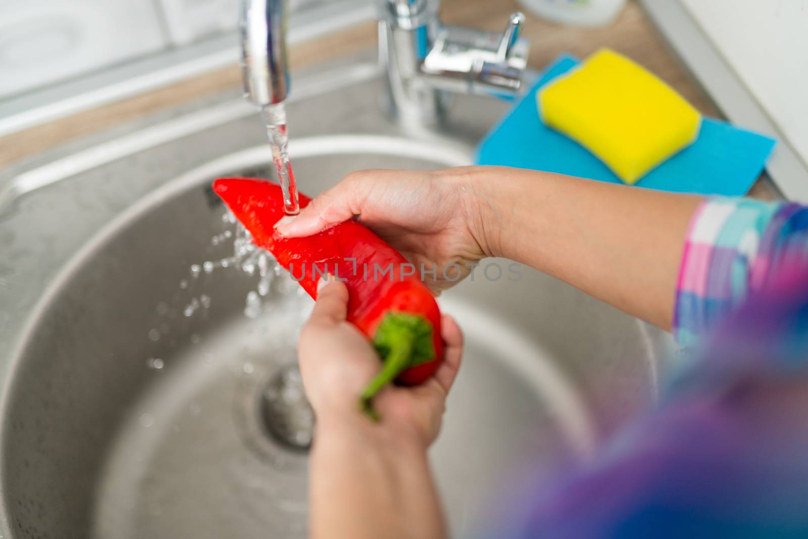 Close-up of a woman hands washing fresh red pepper in the kitchen. Rear view. Unrecognizable person.