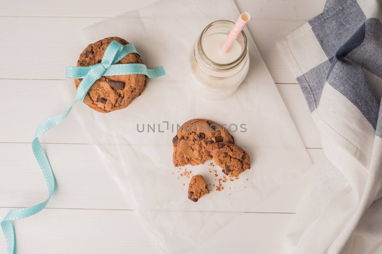 Chocolate chip cookies with a blue ribbon and a glass of milk with a straw on a white wooden table background. Vintage look.