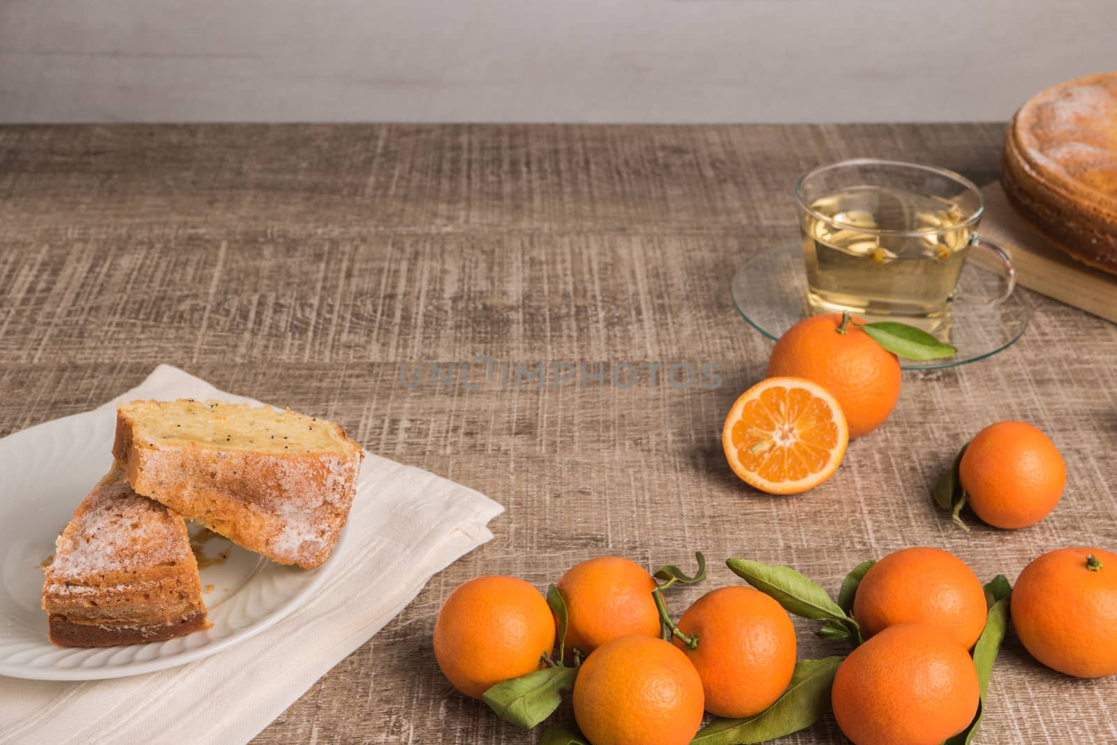 Slices of clementine cake with powdered sugar topping and cup of chamomile tea. Cake on a plate with fresh clementines on wooden board.