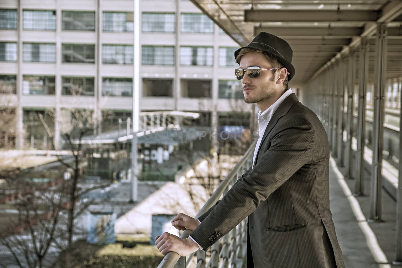 Head and Shoulders Portrait of Stylist Young Man Wearing Suit and Hat Looking to the Side Out Window While Standing on Moving Sidewalk in Building