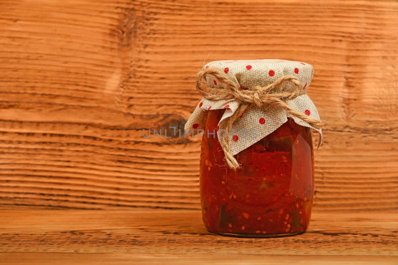 One glass jar of homemade pickled pepper, paprika and eggplant salad with dotted textile top decoration at brown vintage wooden surface