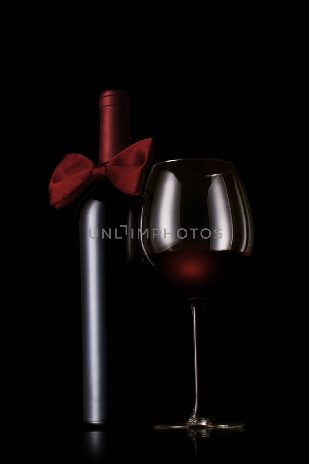 A red wine bottle with bow tie next to a wine glass on black background