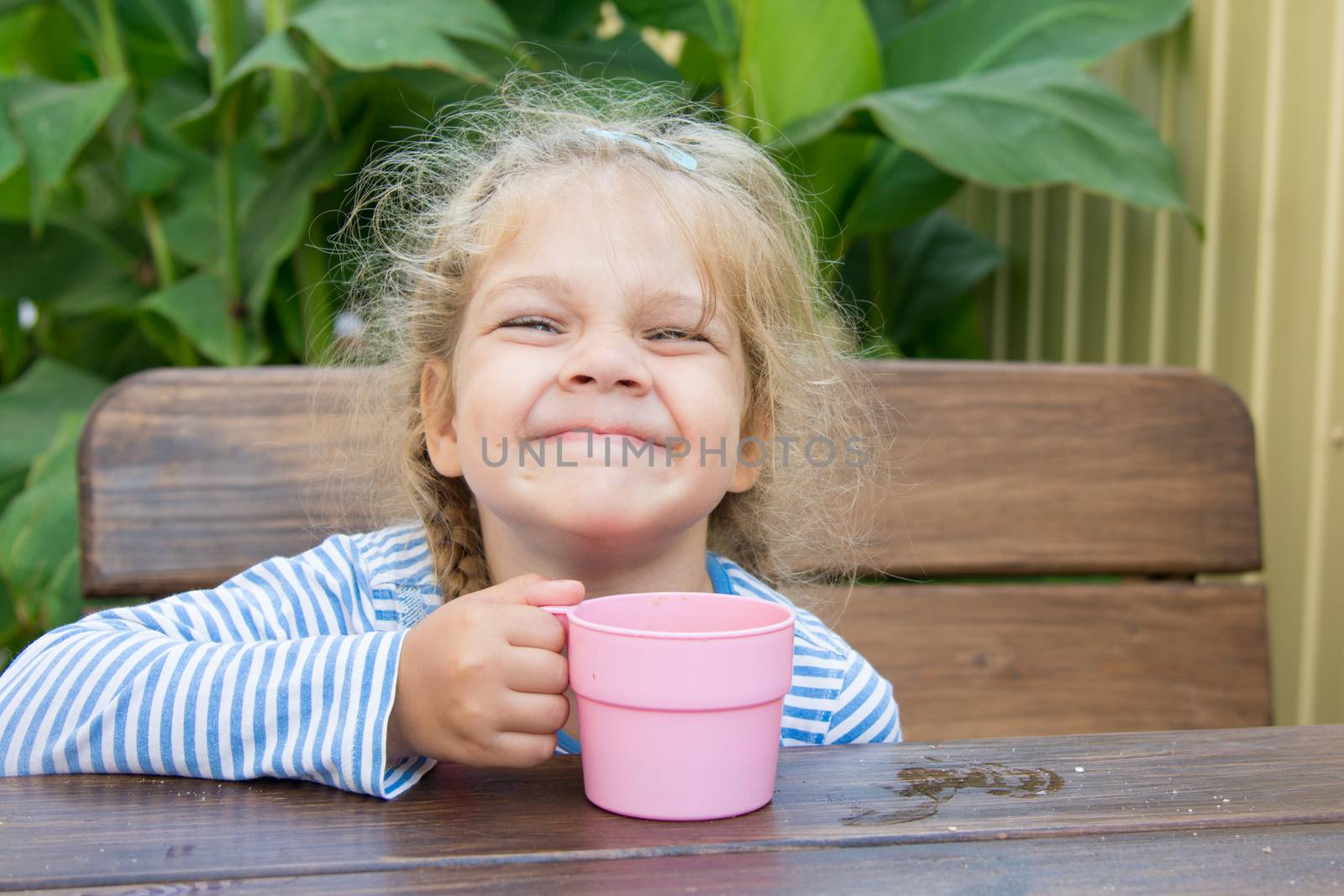Four-year girl sitting at a wooden table on the veranda and drink a cup of pink