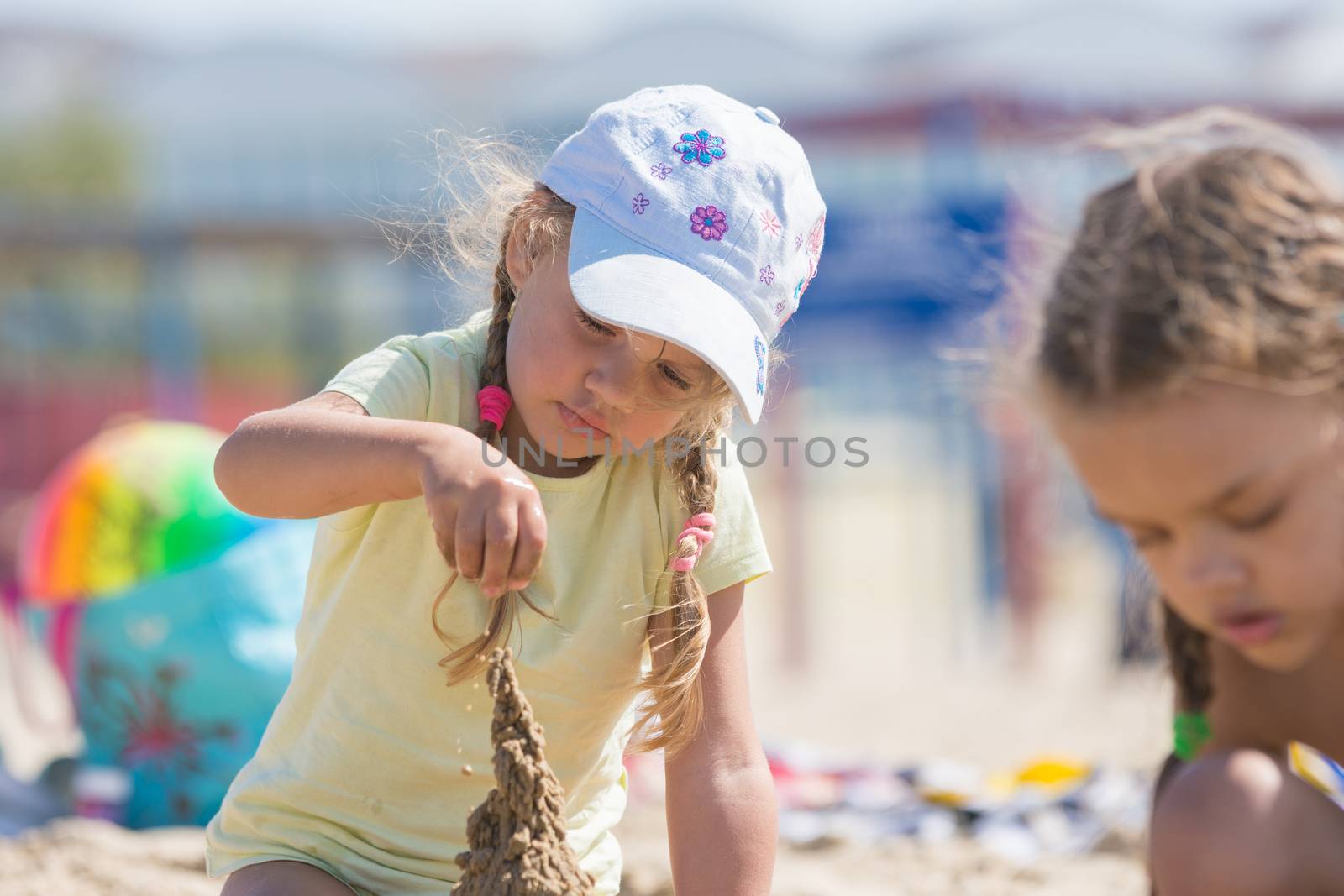 Four-year girl builds a sand castle on the beach of the seaside, beside the other girl sitting