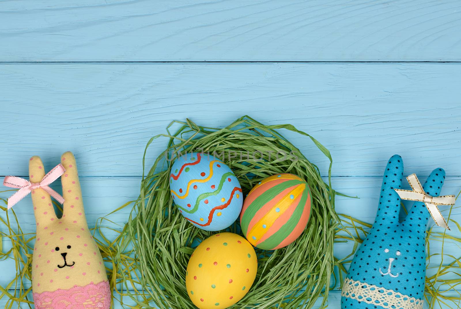 Easter eggs and rabbits. Happy bunny handmade and hand painted multicolored decorated eggs on green straw nest, blue wooden background, copyspace. Unusual creative holiday greeting card 