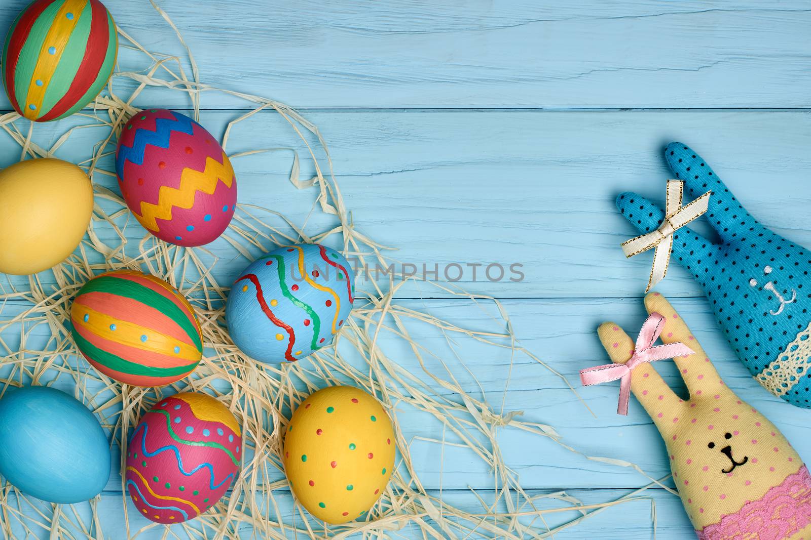 Easter eggs and rabbits. Happy bunny handmade and hand painted multicolored decorated eggs on green straw nest, blue wooden background, copyspace. Unusual creative holiday greeting card 