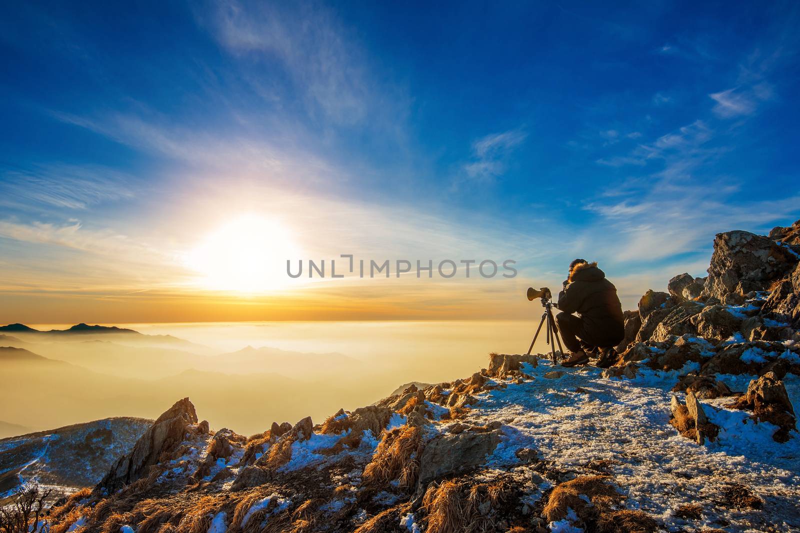 Professional photographer takes photos with camera on tripod on rocky peak at sunset.