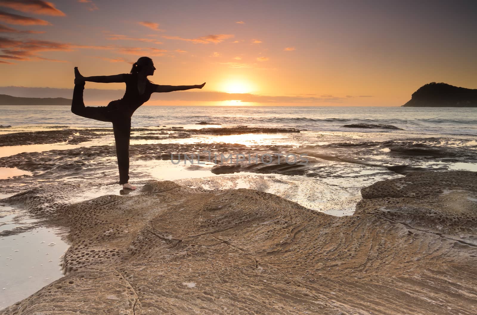A female standing and balancing on one leg practicing a yoga pose, King Dancer Natarajasana;, one of the many asanas to master in yoga.  Behind her the beautiful sunrise, motion of the waves and island add a beautiful backdrop