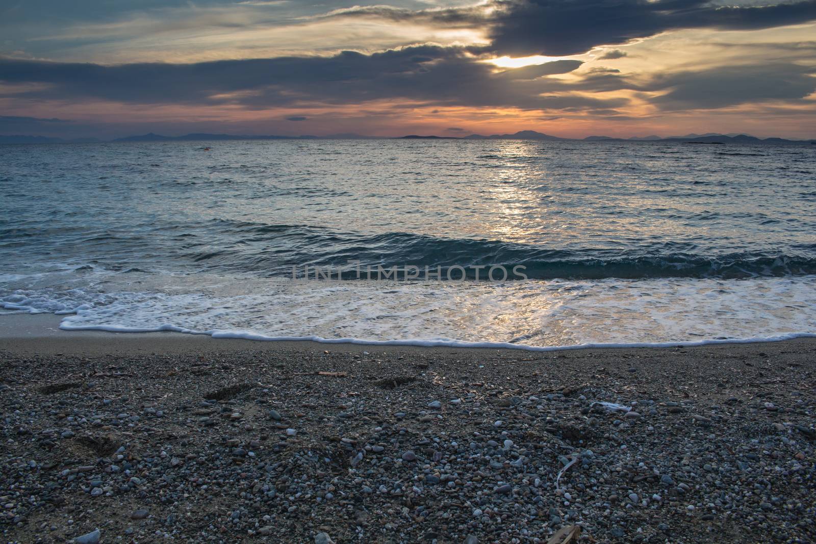 Beach with small stones. Waves with with white edges. Reflection of the sunset on the dark water of Aegean sea. Colorful cloudy sky.