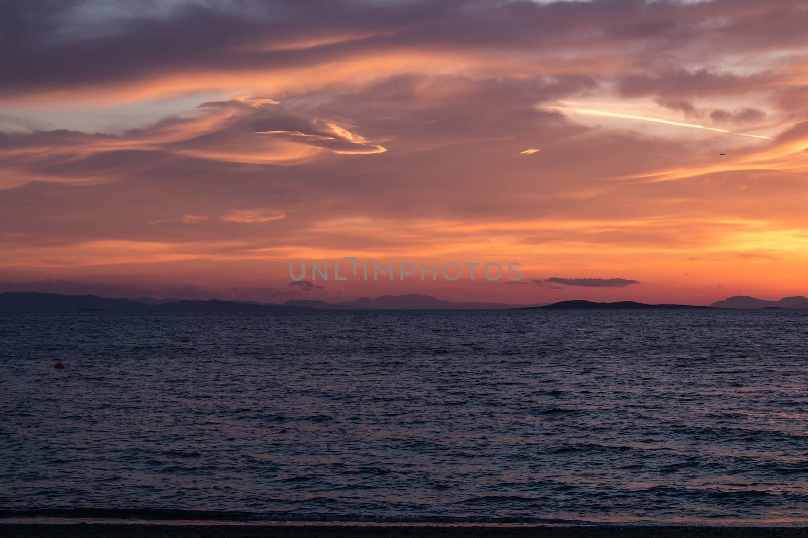 Calm water of the Aegean sea, dark in the early evening. Sunset coloring the sky and clouds in all the shades from yellow to violet.