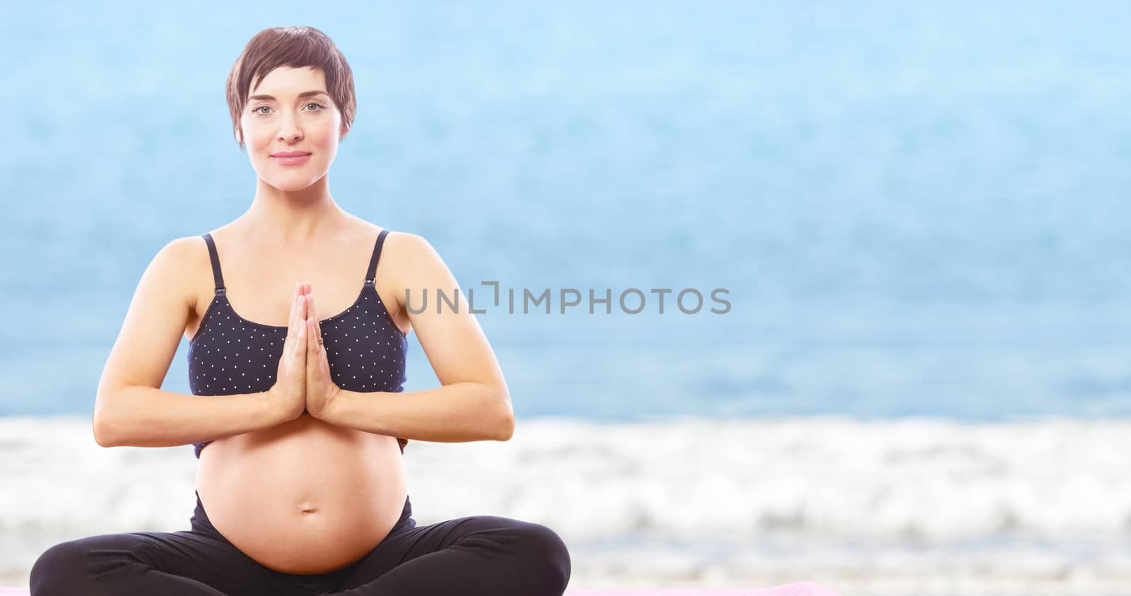 Portrait happy pregnant woman sitting on exercise mat with hands joined against waters edge at the beach