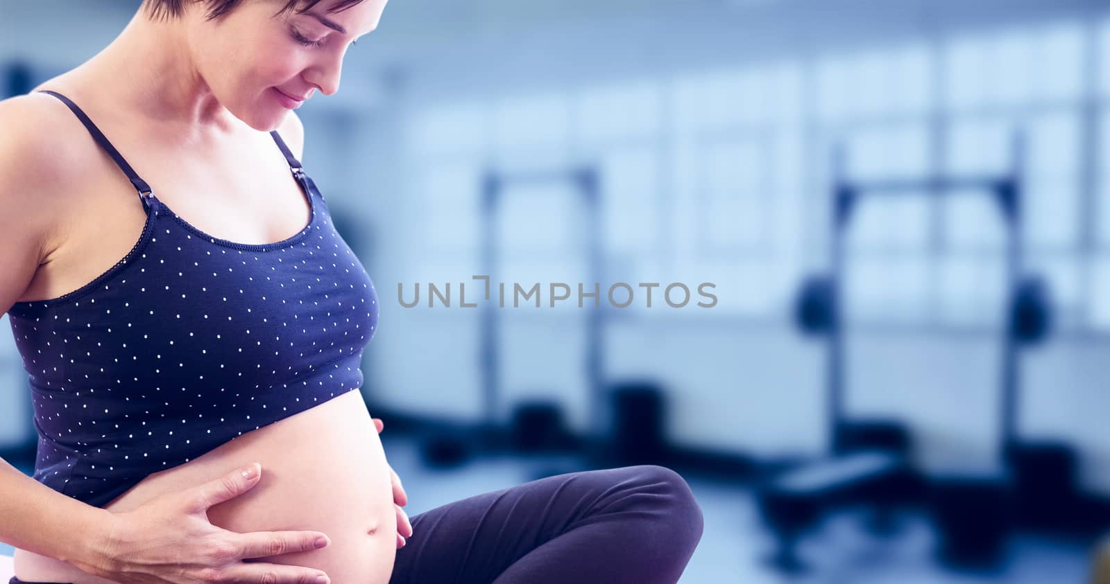 Happy pregnant woman holding belly while sitting on exercise mat against gym