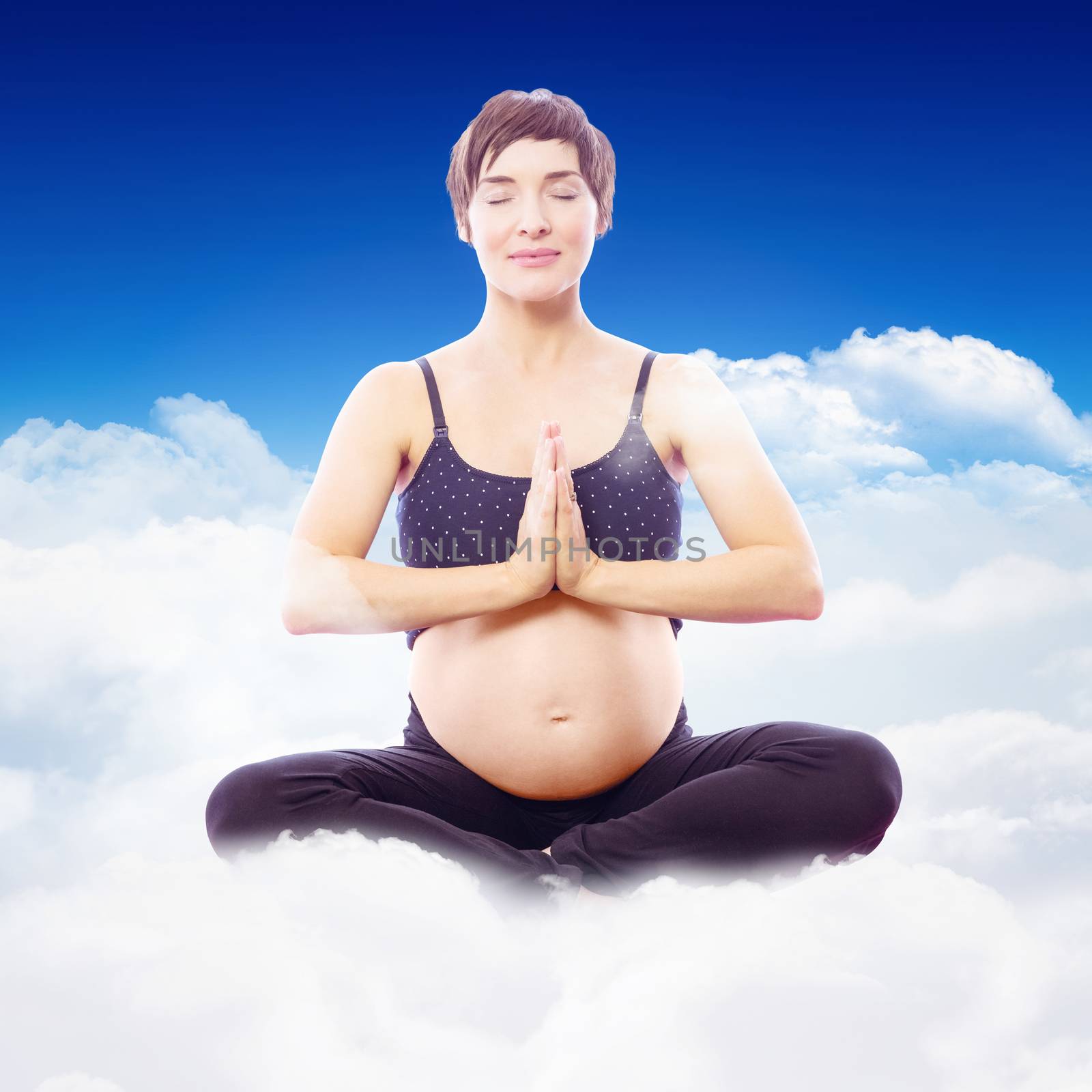 Pregnant woman sitting on exercise mat with hands joined against bright blue sky over clouds