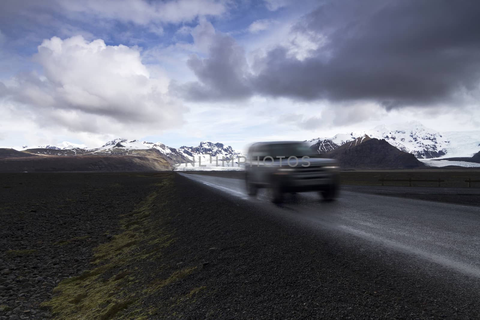 Horizontal photo of an off road car on a straight asphalt road coming from the mountains with clouds above and the Vatnajokull glacier in the background, Iceland