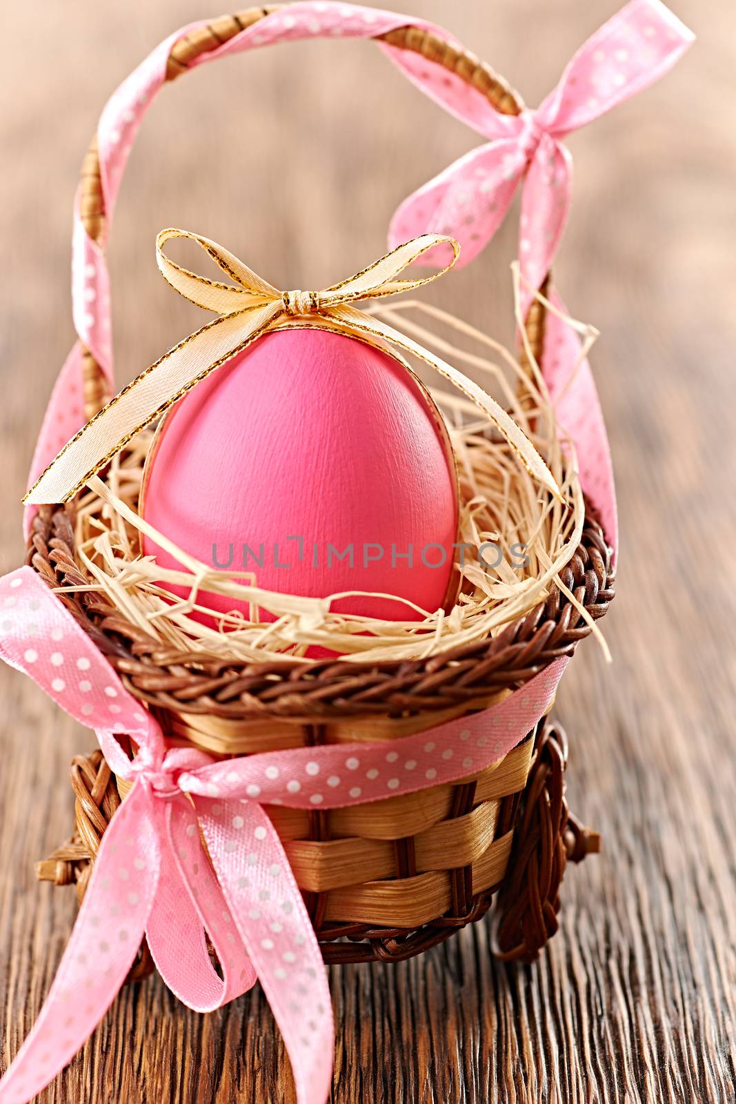 Easter egg in basket. Hand painted decorated pink egg with bow on wooden background. Unusual creative holiday greeting card 