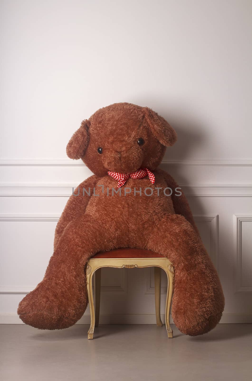 Huge toy bear with a dotted bow standing on a chair against a white wall