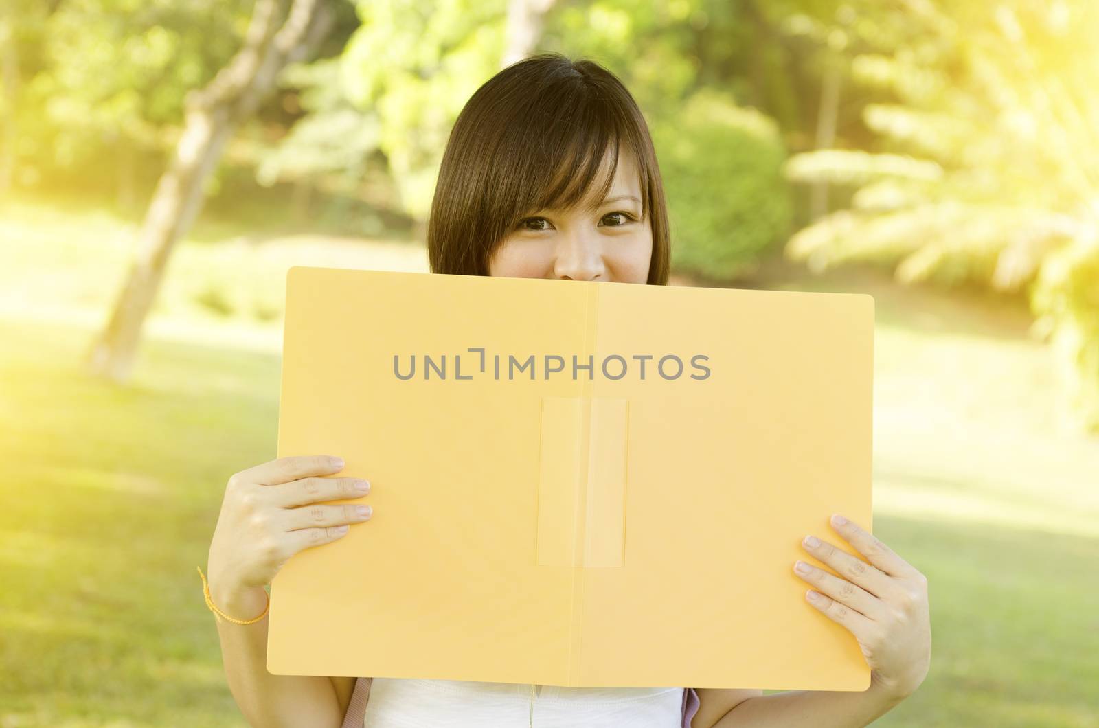 Young Asian college girl student standing on campus lawn, smiling and covered face by file folder.