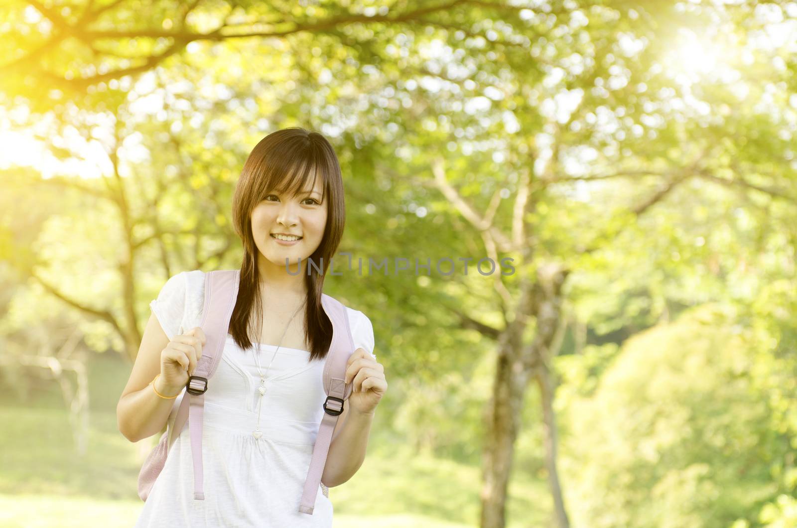 Young Asian teen student standing on campus lawn, with backpack and smiling.