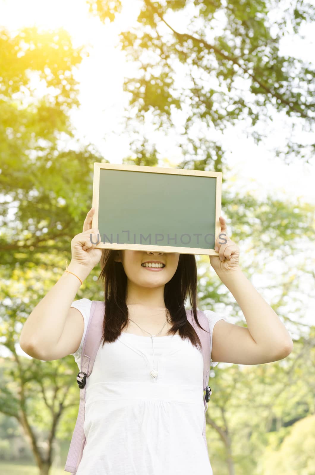 Young Asian college girl student standing on campus lawn, hands holding a blank chalkboard covering face and smiling.