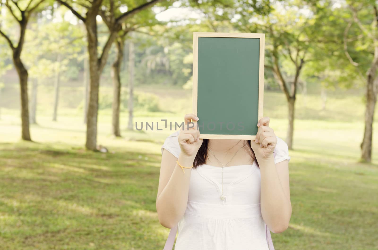 Young Asian college girl student standing on campus lawn, hands holding a blank chalkboard and covered face.
