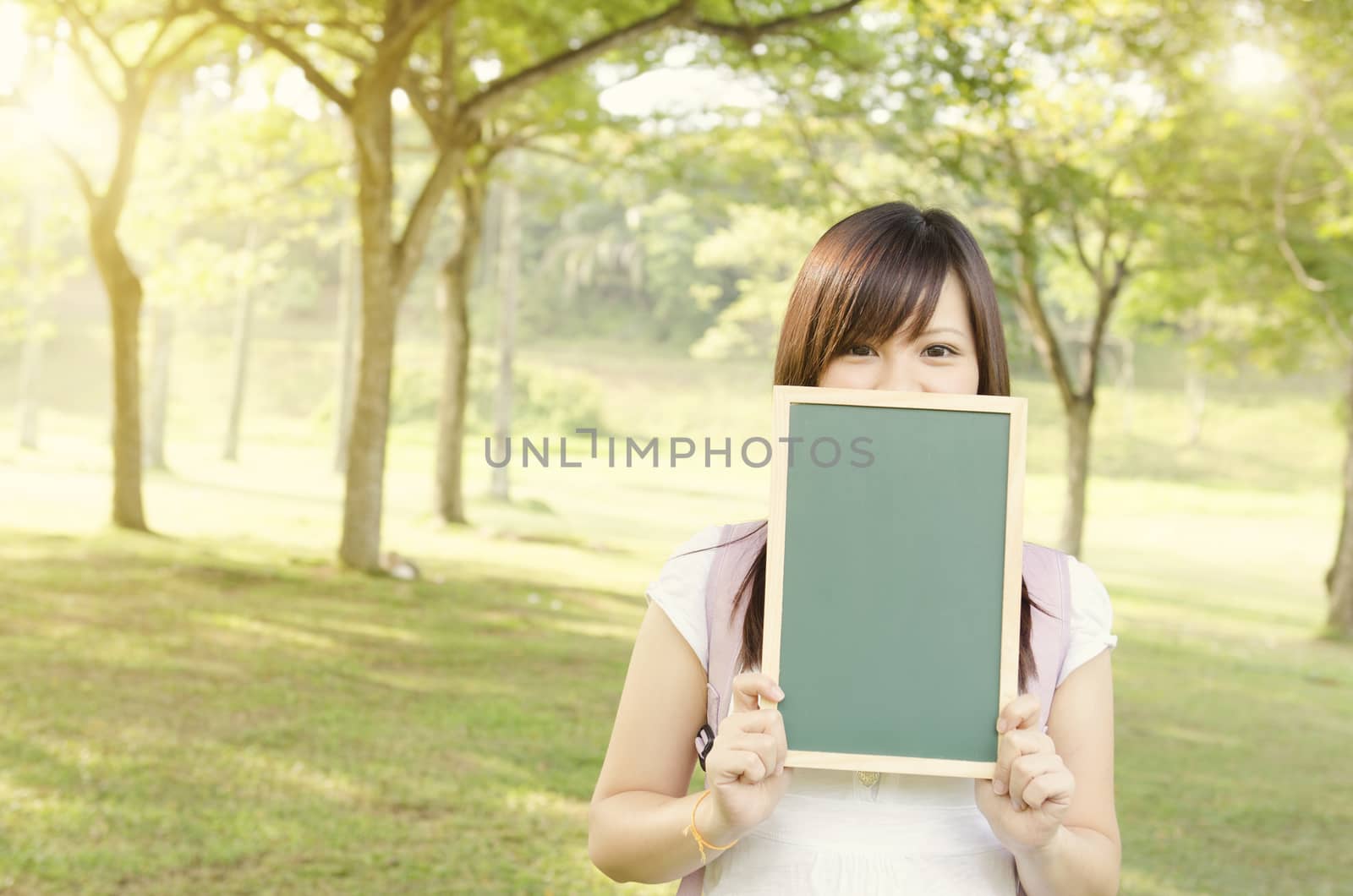 Young Asian college girl student standing on campus lawn, hands holding a blank chalkboard and covered mouth.