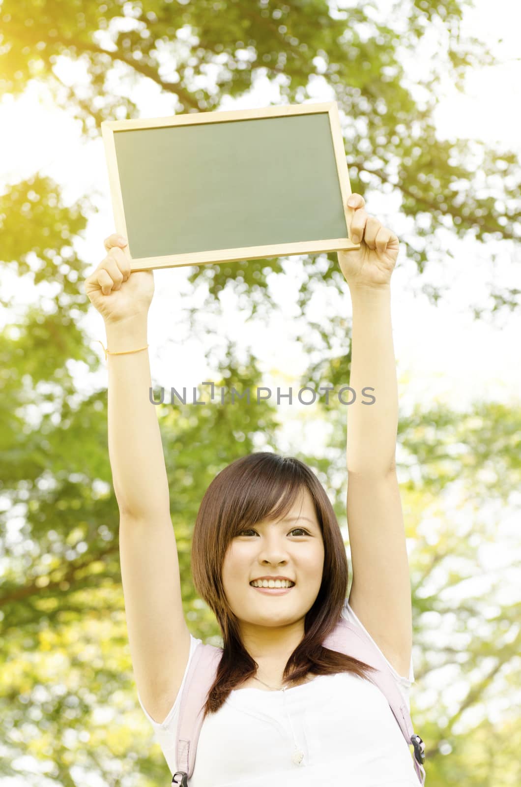 Young Asian college girl student standing on campus lawn, hands raised a blank chalkboard and smiling.