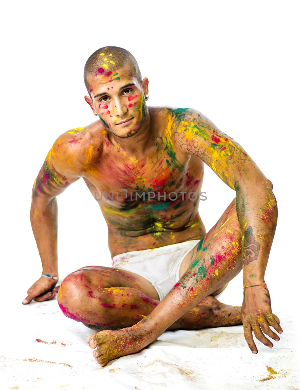 Attractive young man shirtless, skin painted all over with bright Holi colors, looking at camera, sitting, isolated on white background