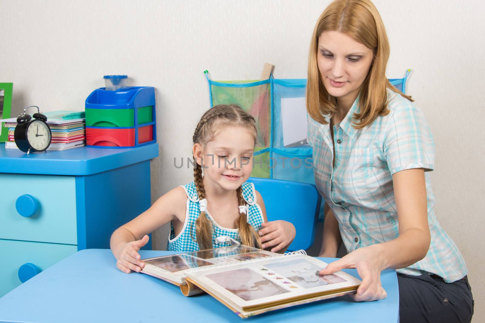 Adult young beautiful girl shows a five-year girl in the photo album sitting at the children table at home