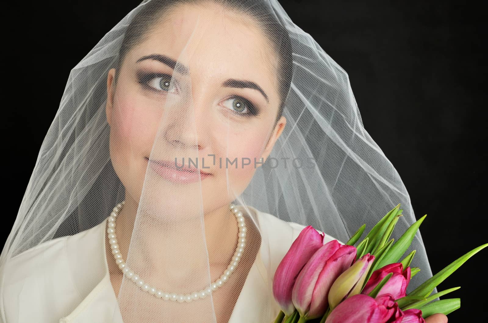Female model, face covered with veil. Bride holds tulips bouquet. Portrait in studio with black background.
