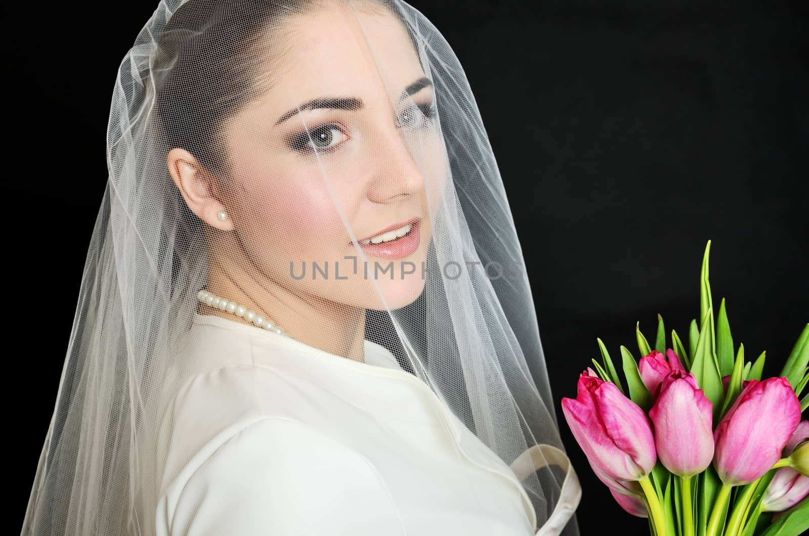 Female model, face covered with veil. Bride holds tulips bouquet. Portrait in studio with black background.
