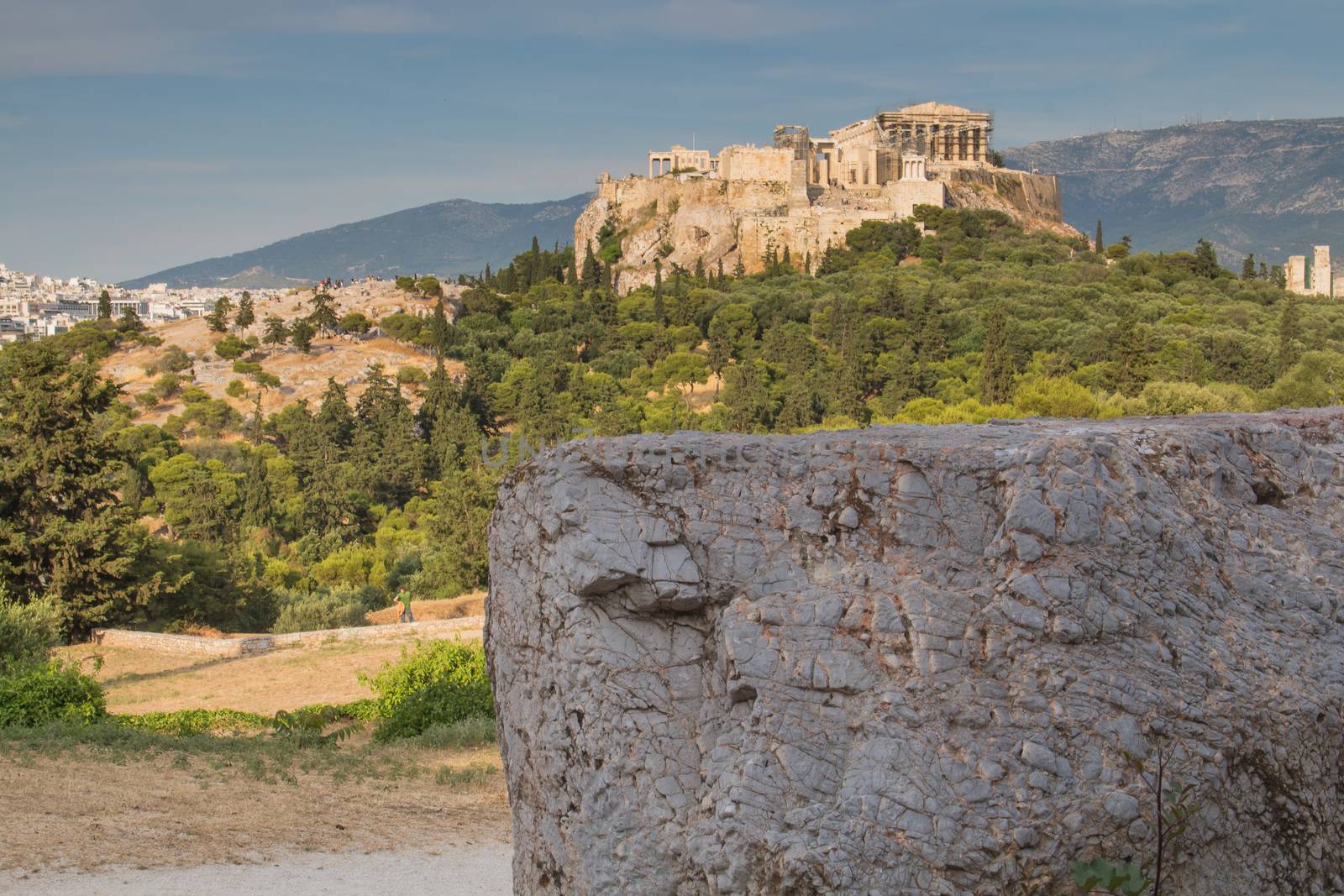 View from a park on the important greek monument: Acropolis, built on the other hill. Big piece of marble in the foreground. Cloudy early evening sky.