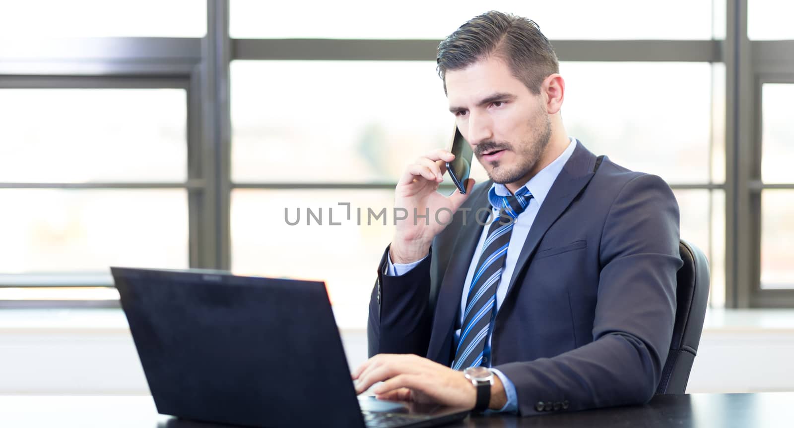 Portrait of successful corporate businessman in bright modern office focused on data on his laptop computer while talking on mobile phone. Business and entrepreneurship concept.