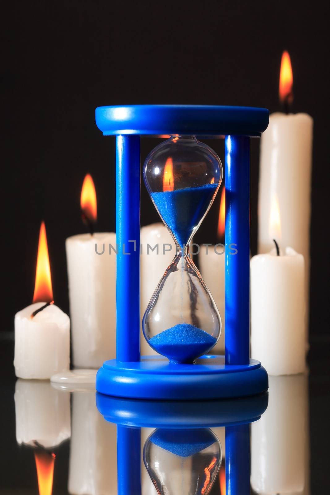 Blue hourglass against set of lighting candles on black background