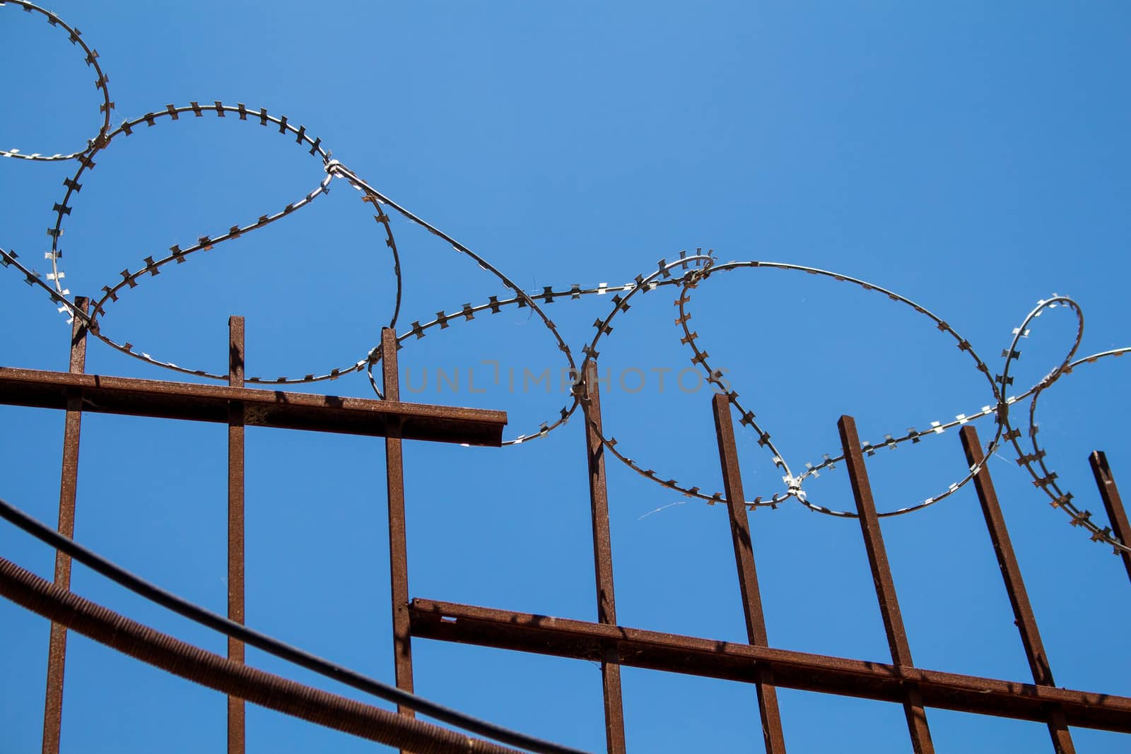 Enlightened barbed wire on the top of a metal fence. Bright blue sky in the background.