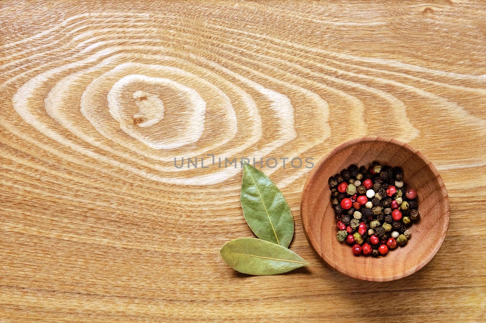 Fragrant colorful pepper in a wooden bowl and two dried bay leaves on a light wooden table. Top view.