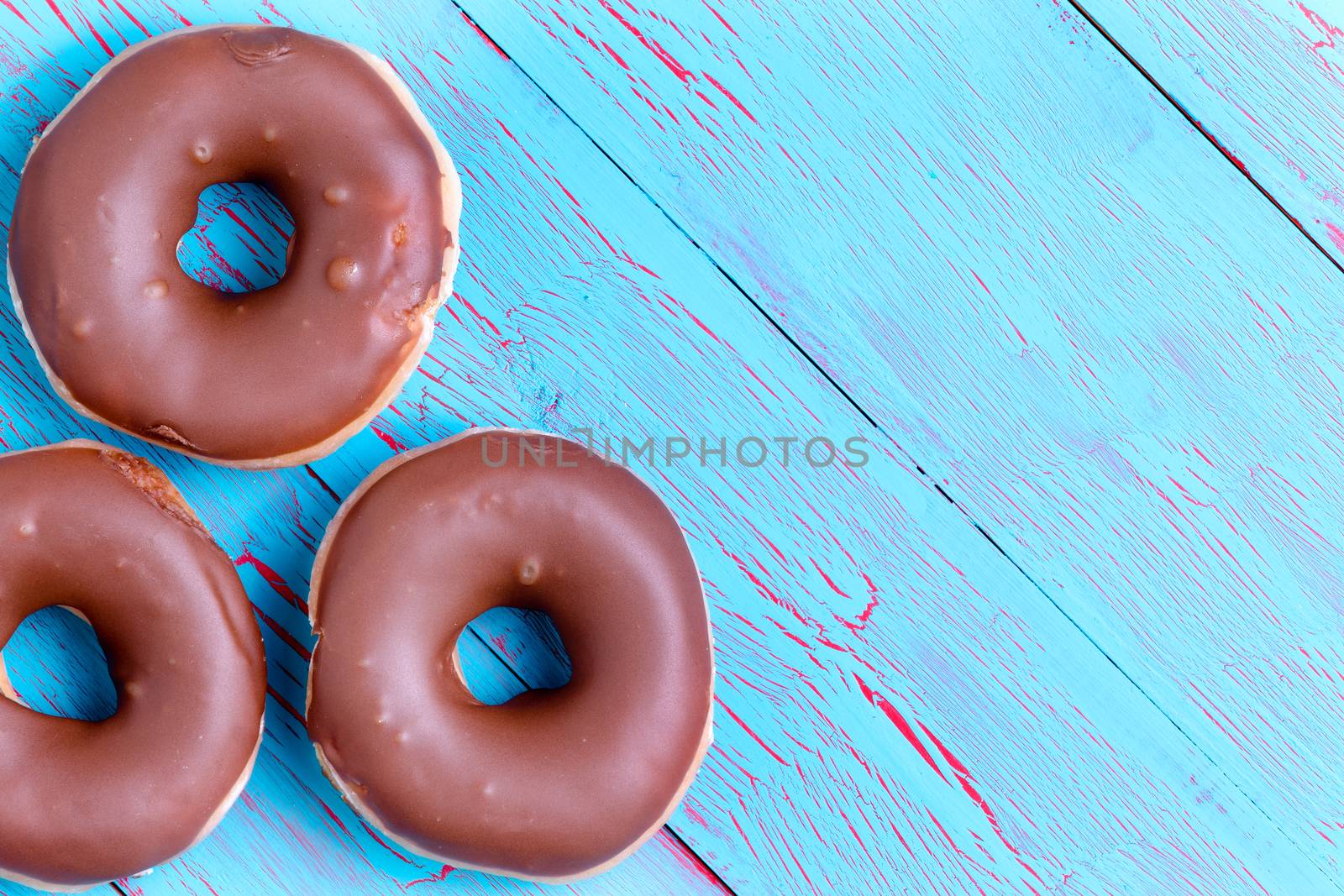 Three crispy cream filled chocolate ring donuts topped with chocolate icing on a rustic turquoise blue wooden background with copy space
