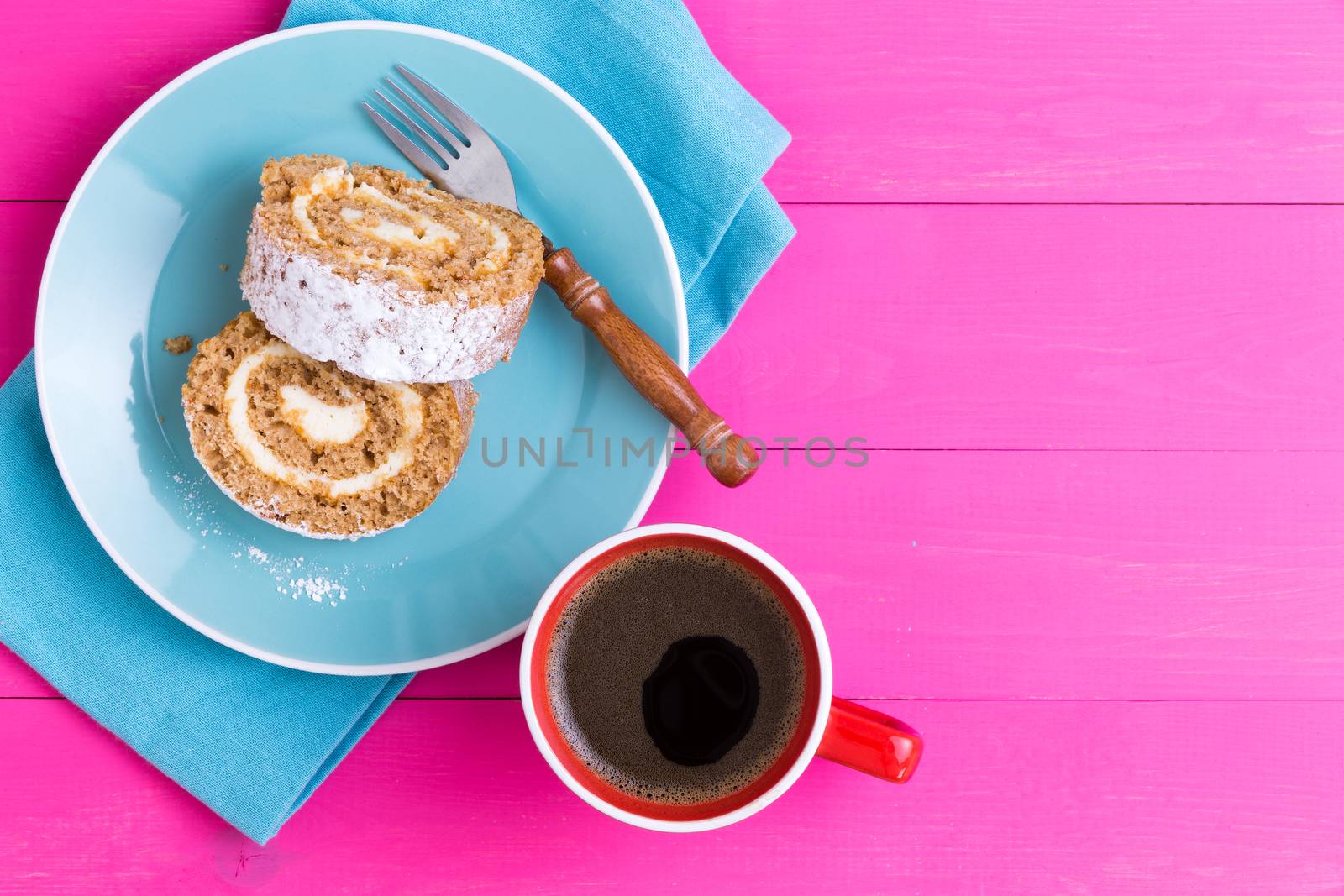 First person view of two pieces of roulo cake dessert and mug of coffee next to fork on napkin over pink wooden table