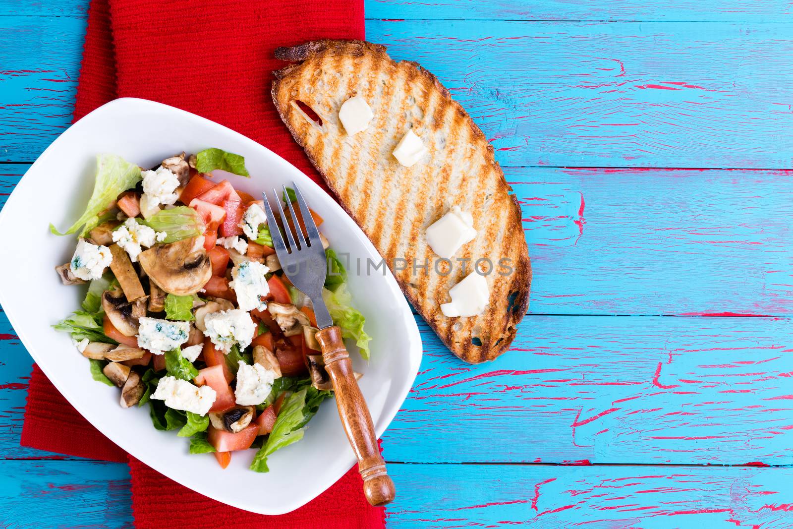 Healthy mixed salad with feta cheese, lettuce and tomato served on a colorful blue weathered picnic table with a red napkin and toast topped with butter, overhead view with copy space