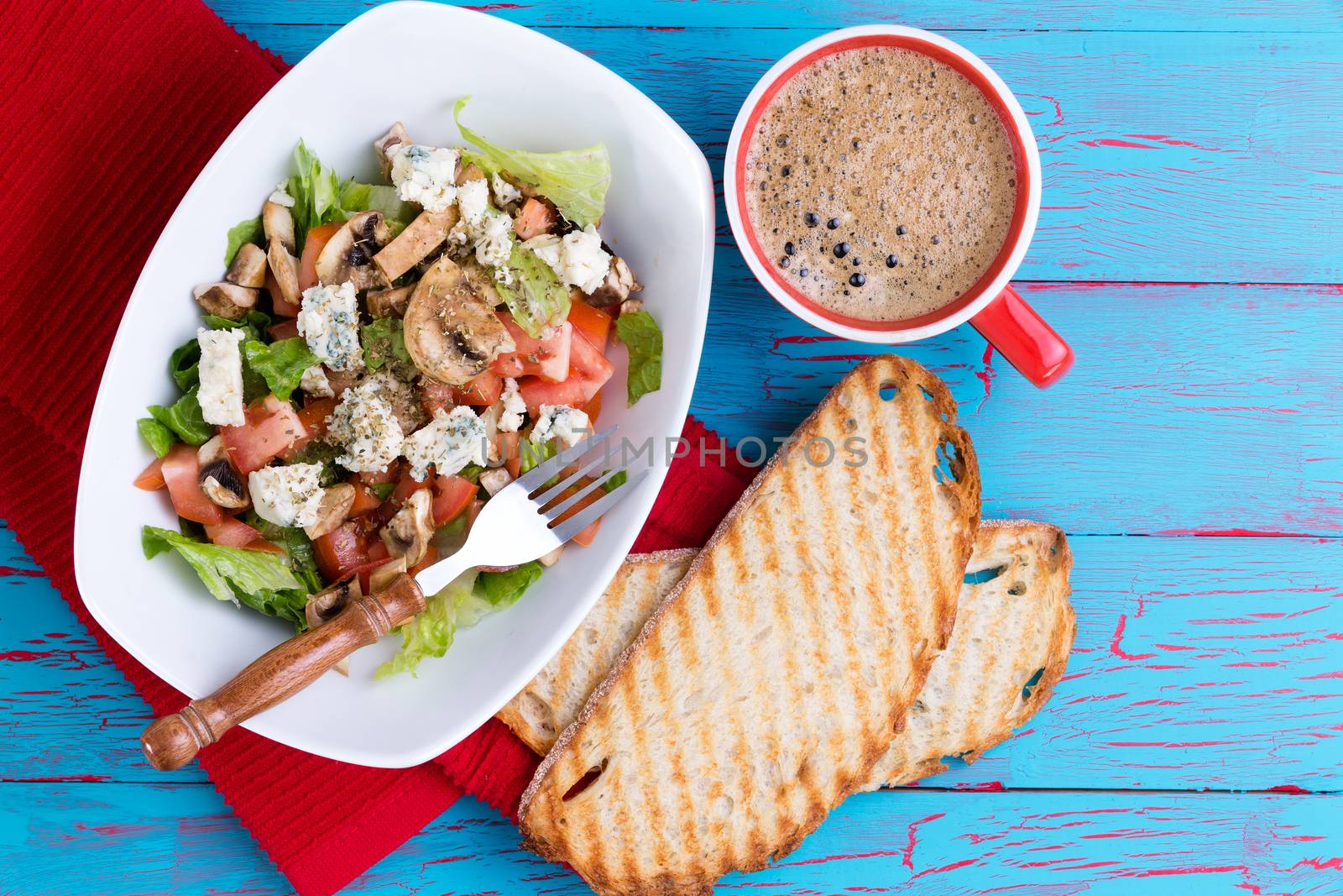 Fresh mixed feta salad on a platter served with frothy espresso coffee and toasted baguette on a colorful tropical blue picnic table with copy space viewed from above