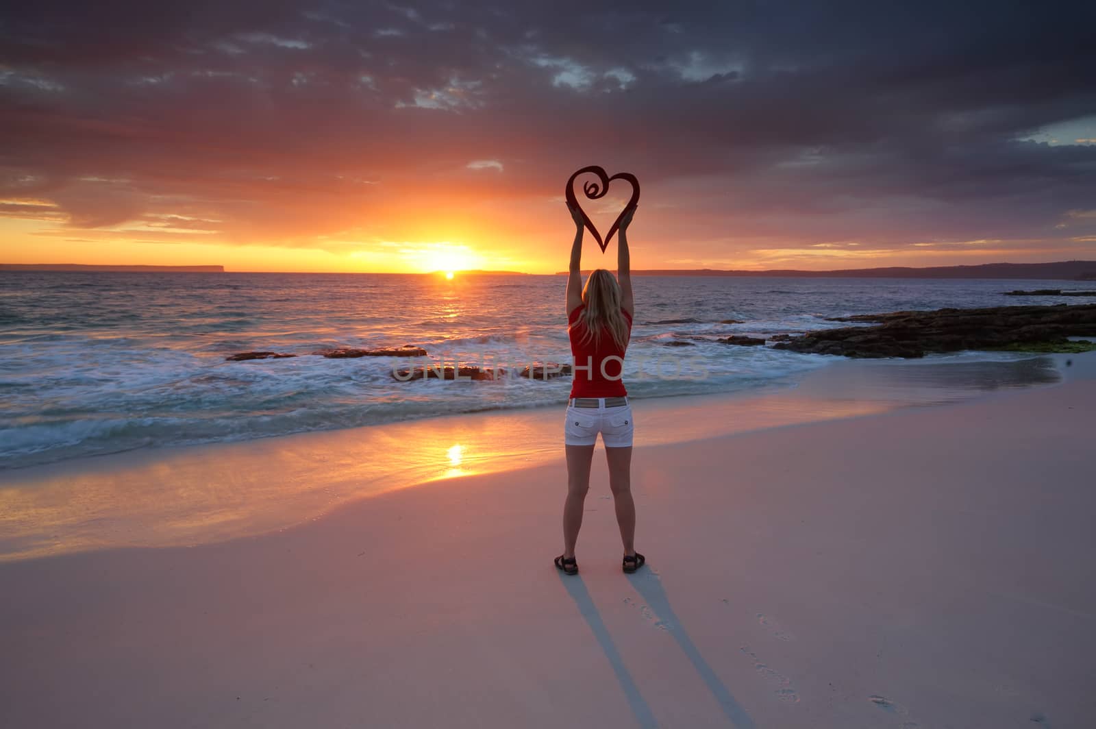 Love Jervis Bay Australia ezpecially when the sunrise is as good as this one casting a red orange glow into the sky and across the water and the usual white sands giving everything a red glow.  Woman holds a red heart above her head