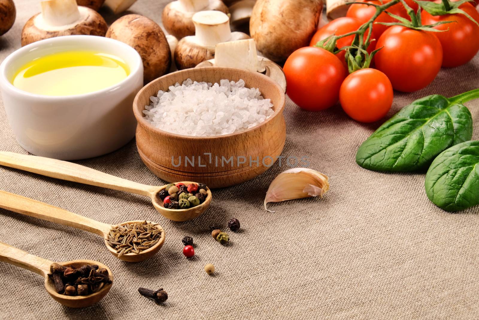 Various spices in small wooden spoons, salt, vegetable oil, cherry tomatoes, mushrooms, spinach and garlic on a linen fabric.