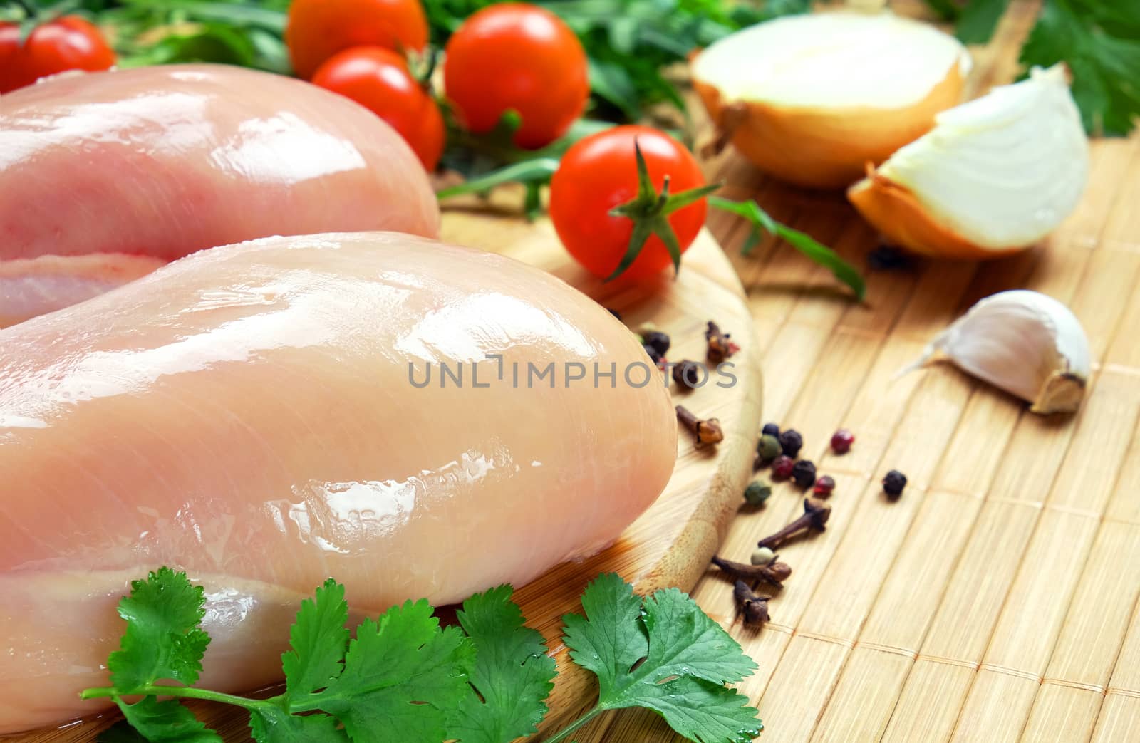 Raw chicken fillet on a round bamboo cutting board, cherry tomatoes, cilantro, onion,  garlic and pepper.