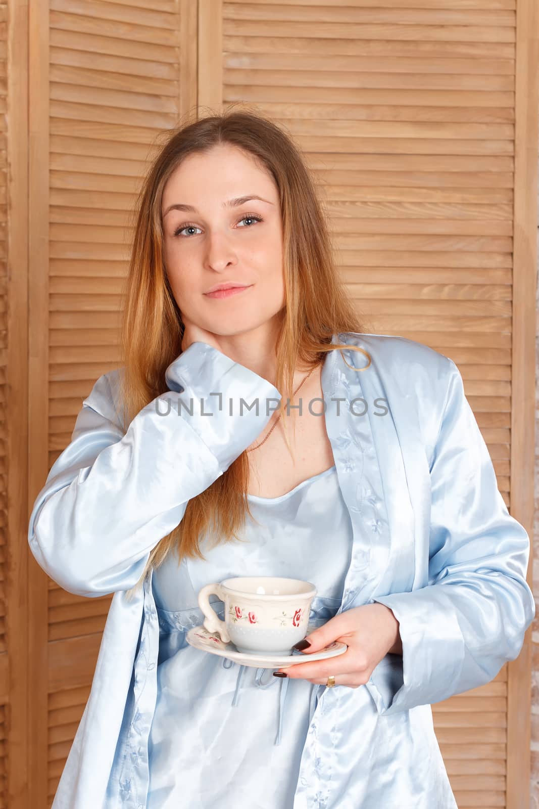 The  yawning woman is standing near window and holding cup
