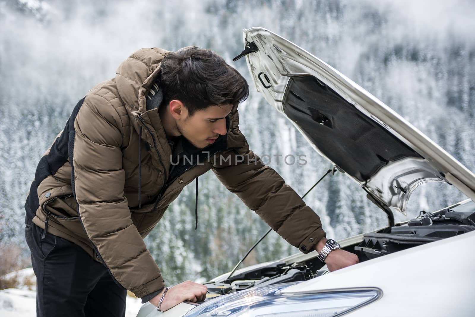 Young man near car with open hood inspecting engine. Snowy forest on background