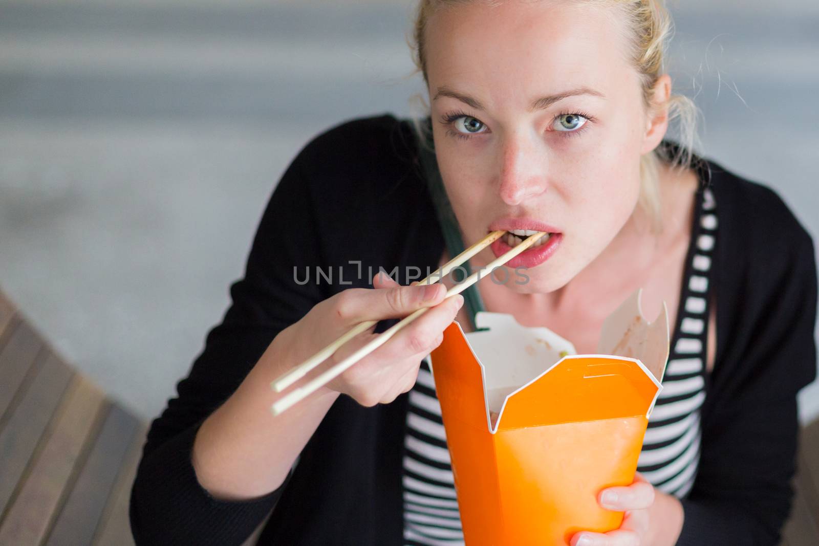 Beautiful young woman, sitting on the bench in park, holding a fast food lunch box, eating noodles from Chinese take-away with traditional wooden chopsticks.