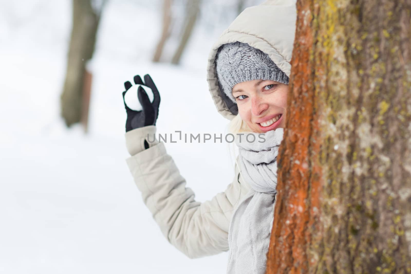 Cute casual young woman hiding behind tree while snowball fighting in winter time.