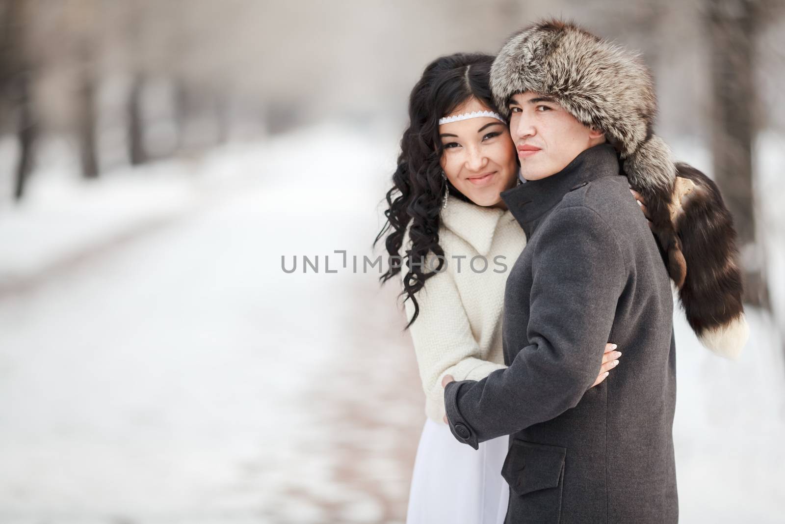 Beautiful wedding couple, exotic asian bride and groom embraced. Young man in winter coat and fur hat, bride in white wedding dress with sheepskin veil. Cold season warm clothing, copy space for text.