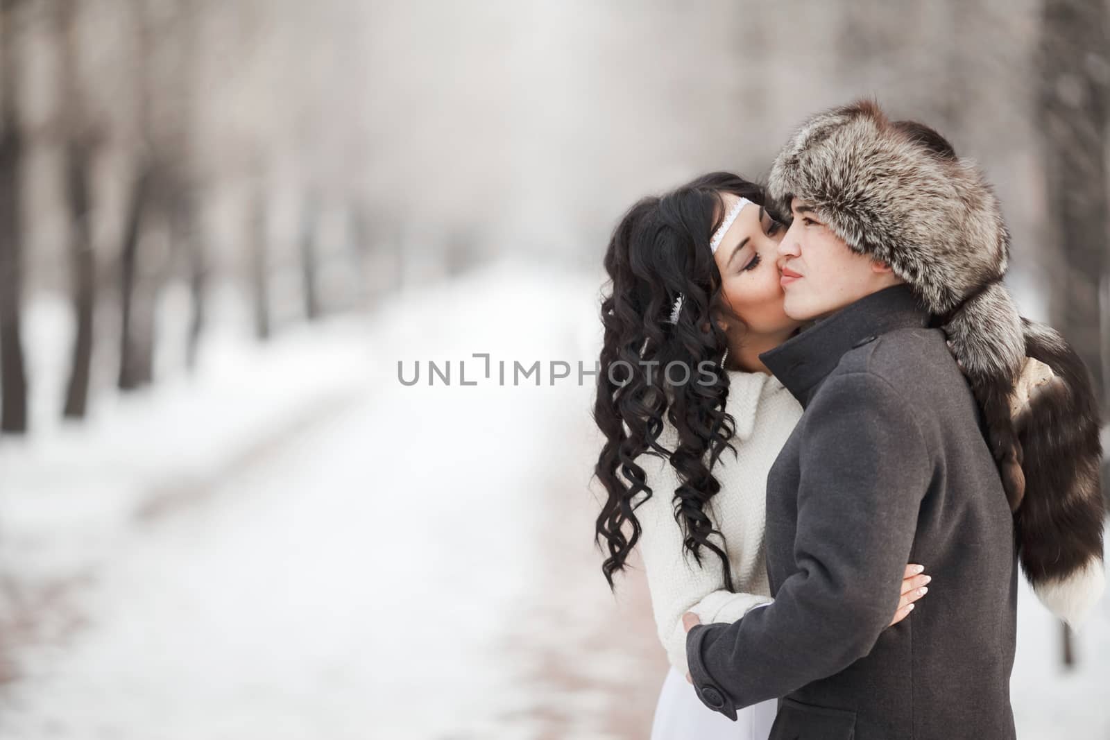 Exotic asian bride and groom kissing in middle of snowy winter alley. Young man in winter coat and fur hat, bride in white wedding dress with sheepskin. Cold season warm clothing. Copy space for text.