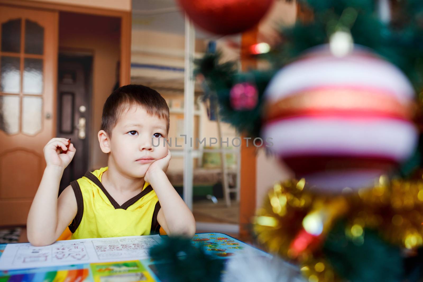 Dreamy little boy sitting on a background Christmas tree writes his wish list on a piece of paper and dreaming about gifts from Santa Claus. He is preparing for the Christmas and New Year holidays.